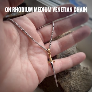 May include: A silver sword pendant on a rhodium medium Venetian chain necklace. The sword is gold-toned with a pointed tip and a decorative handle.