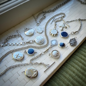 May include: A collection of antique silver necklaces and pendants. The pendants feature various designs, including crosses, floral patterns, and geometric shapes. Some pendants have a dark blue center. The necklaces are displayed on a white surface.