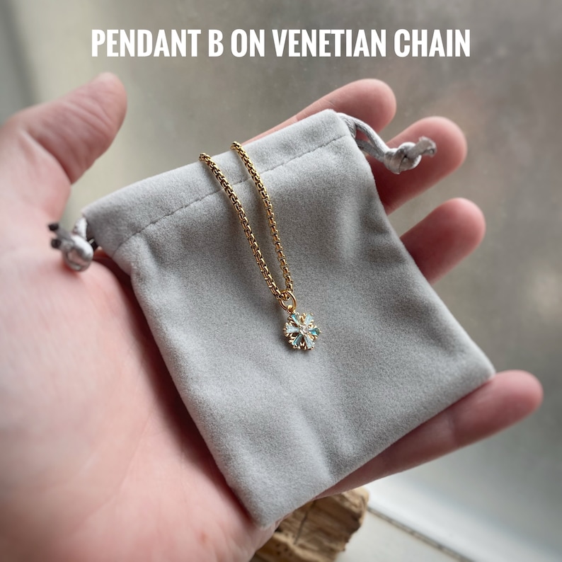 K&ouml;nnte beinhalten: Eine goldene Kettenkette mit einem Schneeflockenanh&auml;nger mit blauen und wei&szlig;en Akzenten. Die Kette wird in einem grauen Samtbeutel pr&auml;sentiert.