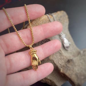 Peut inclure: Un collier doré avec un pendentif en forme de main. Le pendentif présente un poignet et des doigts détaillés. La chaîne est de conception fine et maillée. Un autre collier avec un pendentif en forme de main argentée est visible en arrière-plan.
