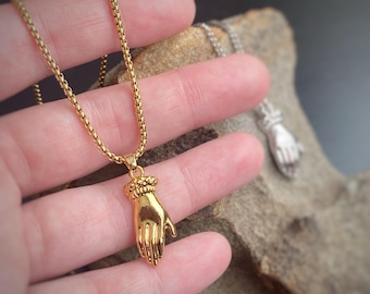 Könnte beinhalten: Eine goldfarbene Halskette mit einem handförmigen Anhänger. Der Anhänger ist mit einem strukturierten Handgelenk und Fingern versehen. Die Kette hat ein feines, gegliedertes Design. Eine weitere silberfarbene Handanhänger-Halskette ist im Hintergrund zu sehen.