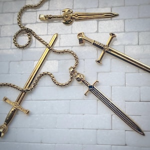 Può includere: Ciondoli a forma di spada color oro e una catena su uno sfondo di mattoni bianchi. I ciondoli variano nel design, uno con una gemma blu. La catena è di colore oro abbinato.