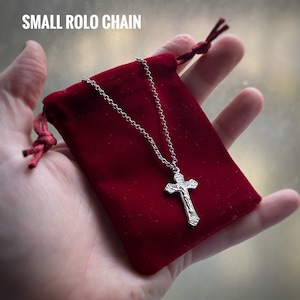 Pu&ograve; includere: Una collana a catena rolo argentata con un ciondolo a croce in argento. La collana &egrave; in una pochette di velluto rosso.