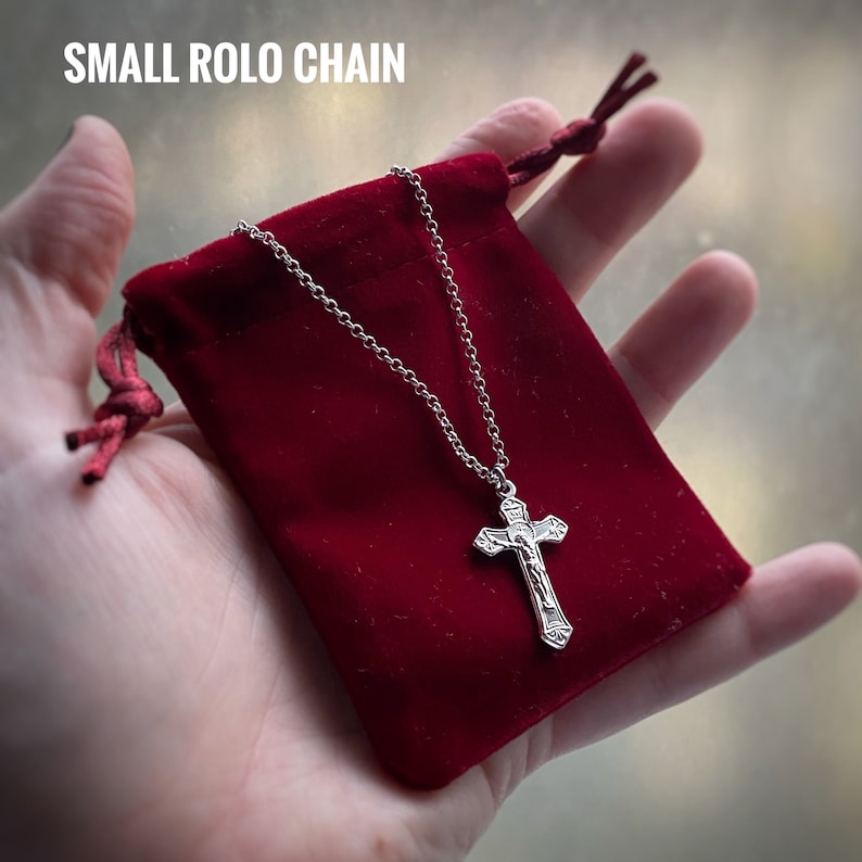 Pu&ograve; includere: Una collana a catena rolo argentata con un ciondolo a croce in argento. La collana &egrave; in una pochette di velluto rosso.