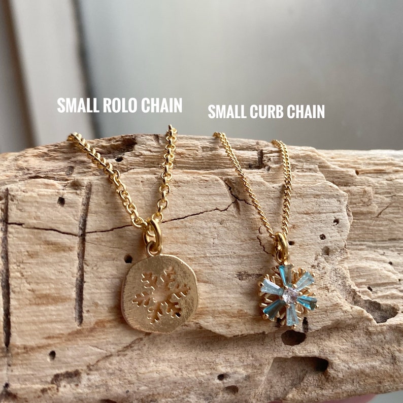 K&ouml;nnte beinhalten: Zwei goldene Halsketten mit Schneeflockenanh&auml;ngern. Die Halskette links hat einen massiven Schneeflockenanh&auml;nger aus Gold. Die Halskette rechts hat einen Schneeflockenanh&auml;nger mit blauen Edelsteinen. Der Text "SMALL ROLLO CHAIN" befindet sich &uuml;ber der linken Halskette und der Text "SMALL CURB CHAIN" &uuml;ber der rechten Halskette.