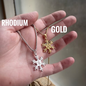 May include: Two snowflake necklaces, one in rhodium and one in gold, on a hand. The necklaces are both chain style with a snowflake pendant.