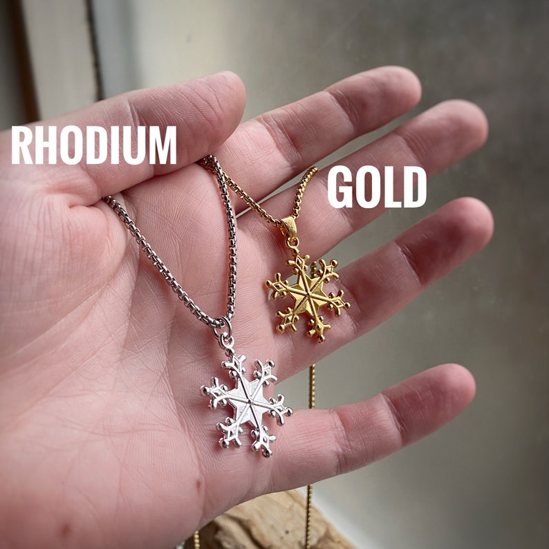 May include: Two snowflake necklaces, one in rhodium and one in gold, on a hand. The necklaces are both chain style with a snowflake pendant.