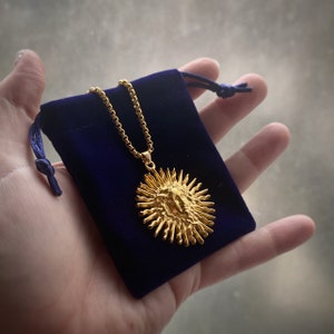 Op de afbeelding: Een goudkleurige zonnehanger ketting met een gedetailleerd gezichtsontwerp, gepresenteerd op een gouden ketting. De ketting wordt gepresenteerd in een donkerblauwe fluwelen zakje met een trekkoordsluiting. De hanger heeft een textuur.