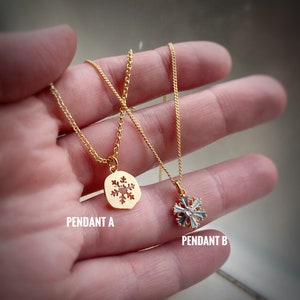 K&ouml;nnte beinhalten: Zwei goldene Halsketten mit Schneeflockenanh&auml;ngern. Anh&auml;nger A ist eine massive goldene Schneeflocke mit einem Ausschnittmuster. Anh&auml;nger B ist eine goldene Schneeflocke mit blauen und wei&szlig;en Kristallen.