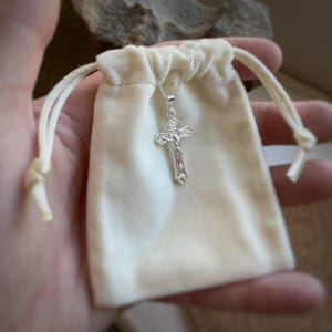 May include: A silver crucifix pendant with a detailed design, resting in a white velvet pouch.