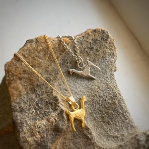 Könnte beinhalten: Zwei Halsketten mit Tieranhängern liegen auf einem strukturierten Stein. Eine Halskette ist goldfarben mit einem Lama-Anhänger. Die andere ist silberfarben mit einem stilisierten Vogelanhänger. Beide Halsketten haben zarte Ketten.