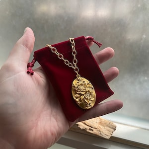 Puede incluir: Un collar de cadena dorada con un gran colgante ovalado que presenta un diseño floral en relieve detallado. El collar se muestra en una bolsa de terciopelo rojo.