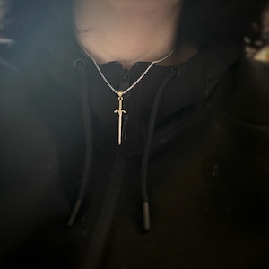 May include: A silver chain necklace with a gold sword pendant. The sword is a simple design with a pointed tip and a cross guard.