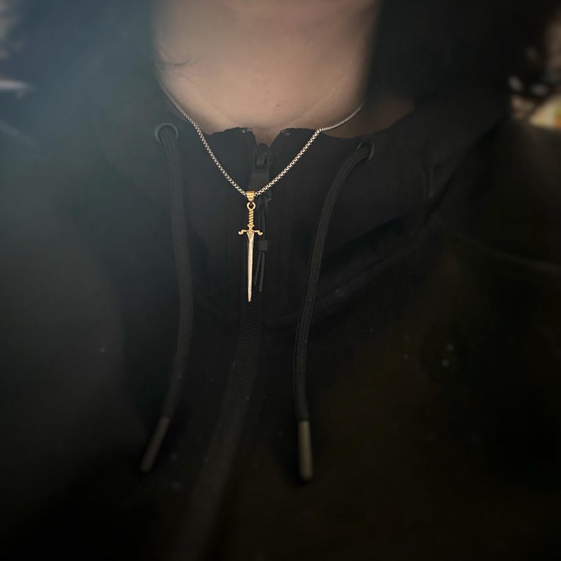 May include: A silver chain necklace with a gold sword pendant. The sword is a simple design with a pointed tip and a cross guard.