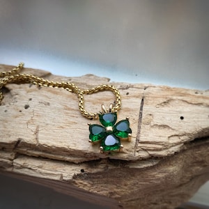 May include: A gold chain necklace with a green four-leaf clover pendant. The pendant is made of gold and has four green gemstones in the shape of hearts.