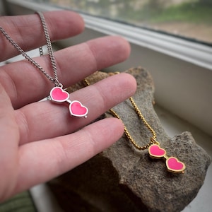 Könnte beinhalten: Zwei Halsketten mit Anhängern in Form von Brillen mit rosa herzförmigen Gläsern. Eine Kette ist silberfarben, die andere goldfarben. Die Schmuckstücke werden auf einer steinernen Oberfläche präsentiert, eine Kette wird in der Hand gehalten.
