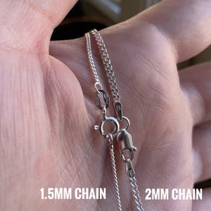 May include: Two silver chains, one 1.5mm and one 2mm, are shown on a person's hand. The chains are both silver and have a clasp.