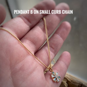 K&ouml;nnte beinhalten: Eine goldene Kettenkette mit einem kleinen, filigranen Schneeflockenanh&auml;nger mit klaren und blauen Kristallen. Der Anh&auml;nger befindet sich an einer kleinen Gliederkette.