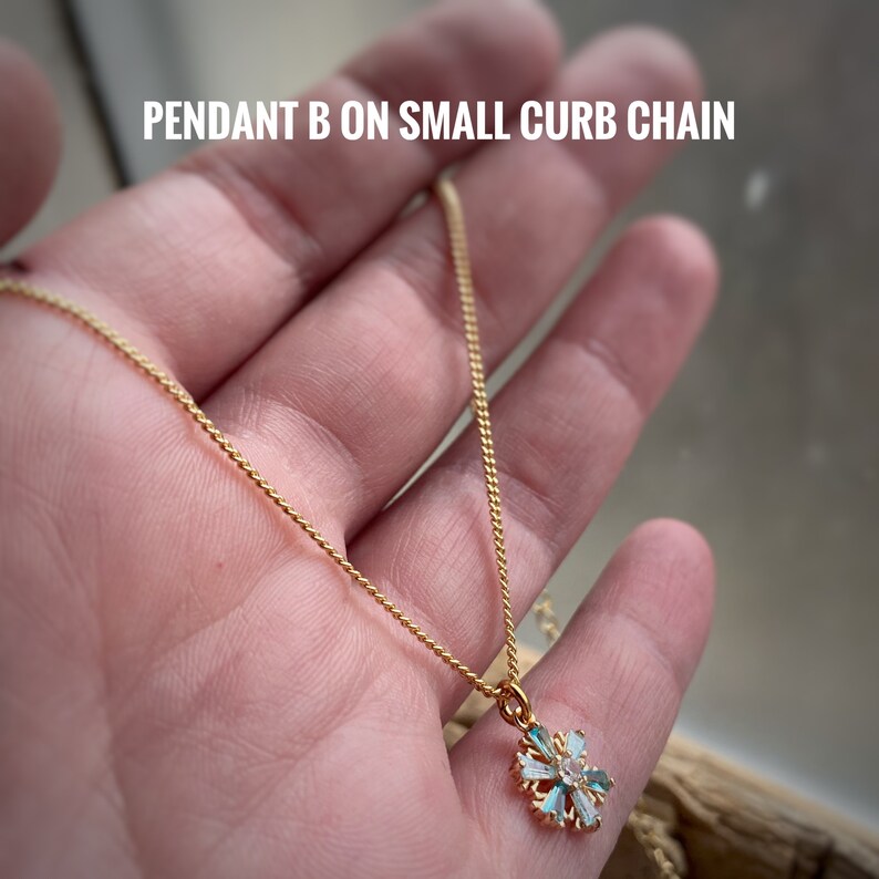 K&ouml;nnte beinhalten: Eine goldene Kettenkette mit einem kleinen, filigranen Schneeflockenanh&auml;nger mit klaren und blauen Kristallen. Der Anh&auml;nger befindet sich an einer kleinen Gliederkette.