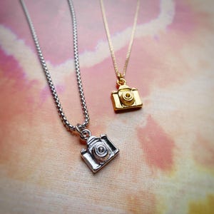 Peut inclure: Deux colliers en chaîne argent avec des breloques pendantes en forme d'appareil photo. Un charme d'appareil photo est en or et l'autre en argent.