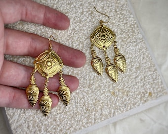 May include: A pair of gold dangle earrings with a round, ornate design and three teardrop-shaped pendants.