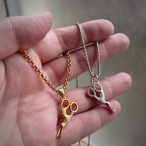 Puede incluir: Dos collares de oro y plata con colgantes de tijeras. El collar de oro tiene un colgante de tijeras detallado con un mango curvo. El collar de plata tiene un colgante de tijeras simple con mangos rectos.