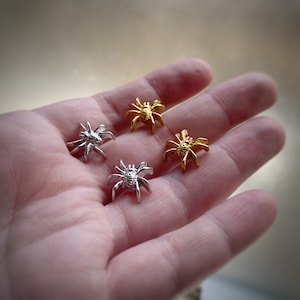 Op de afbeelding: Vier kleine spinnencharms, twee goud en twee zilver, getoond op een hand. De spinnen hebben acht poten en een gedetailleerd lichaam.