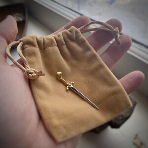 May include: A gold sword pendant with a silver blade, resting in a brown velvet pouch.