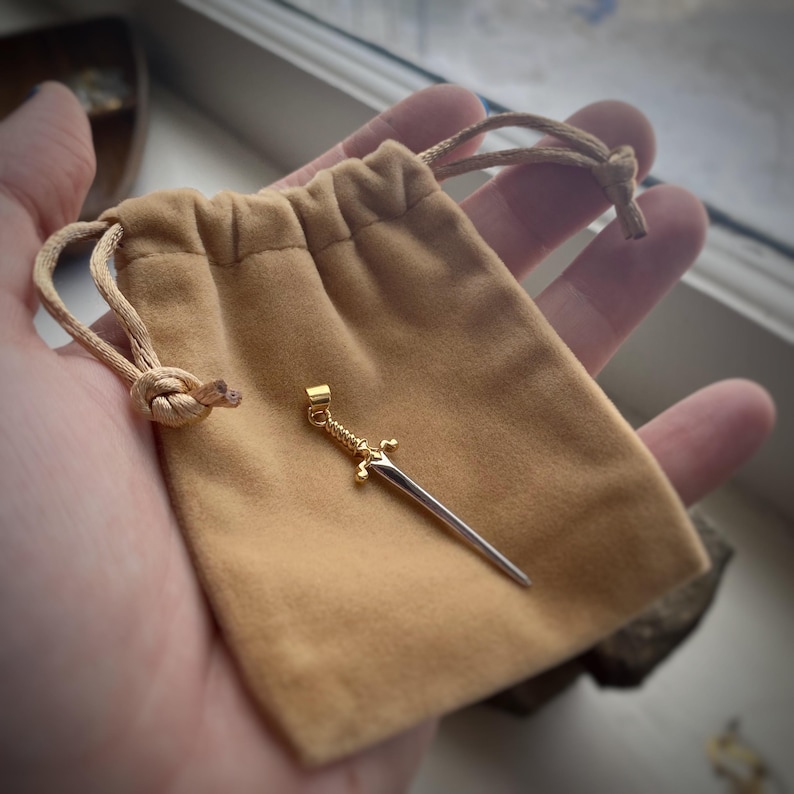 May include: A gold sword pendant with a silver blade, resting in a brown velvet pouch.