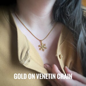 May include: A gold snowflake pendant on a gold chain necklace. The necklace is worn by a person with long dark hair. The text "GOLD ON VENETIN CHAIN" is visible at the bottom of the image.