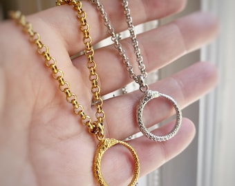 May include: Two necklaces with circular pendants, one gold-toned and one silver-toned, are displayed. The pendants feature a textured snake design, and the chains have a linked design. The necklaces are held up against a soft background.