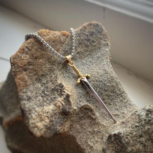 May include: A silver chain necklace with a gold and silver sword pendant. The pendant is detailed with a cross guard and a pointed tip.