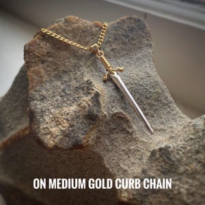 May include: A silver sword pendant on a gold curb chain. The sword has a pointed tip and a decorative handle. The chain is medium in size.