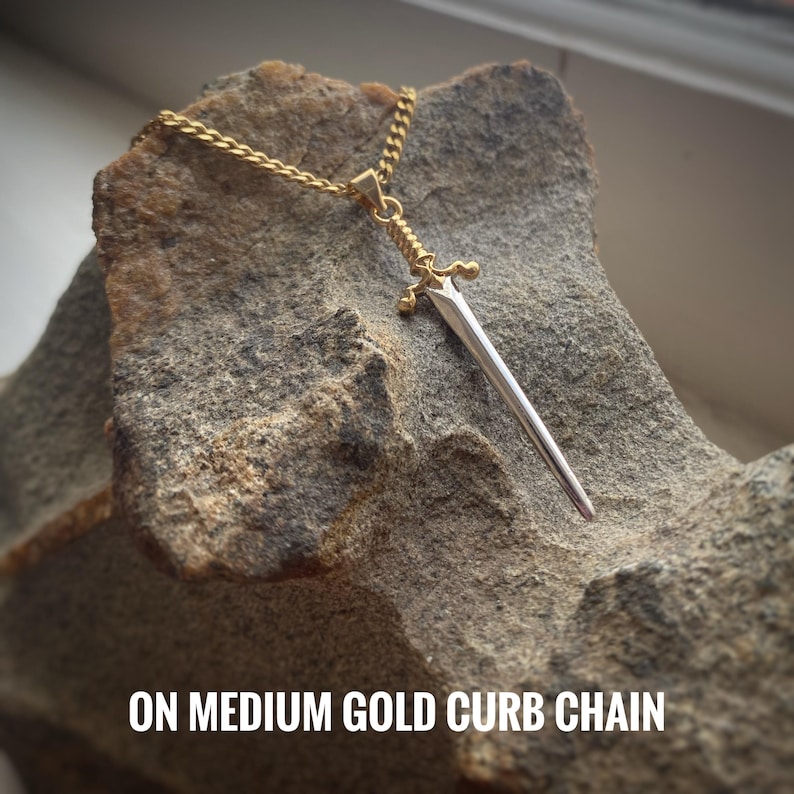 May include: A silver sword pendant on a gold curb chain. The sword has a pointed tip and a decorative handle. The chain is medium in size.