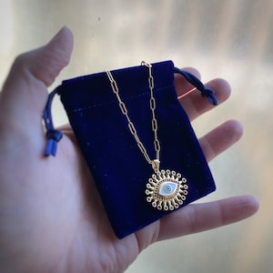 May include: A gold chain necklace with a large evil eye pendant. The pendant is white with a blue eye and surrounded by red gemstones.