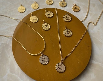 May include: A set of 12 gold zodiac necklaces with a variety of zodiac signs, including Aries, Taurus, Gemini, Cancer, Leo, Virgo, Libra, Scorpio, Sagittarius, Capricorn, Aquarius, and Pisces. The necklaces are displayed on a brown surface.