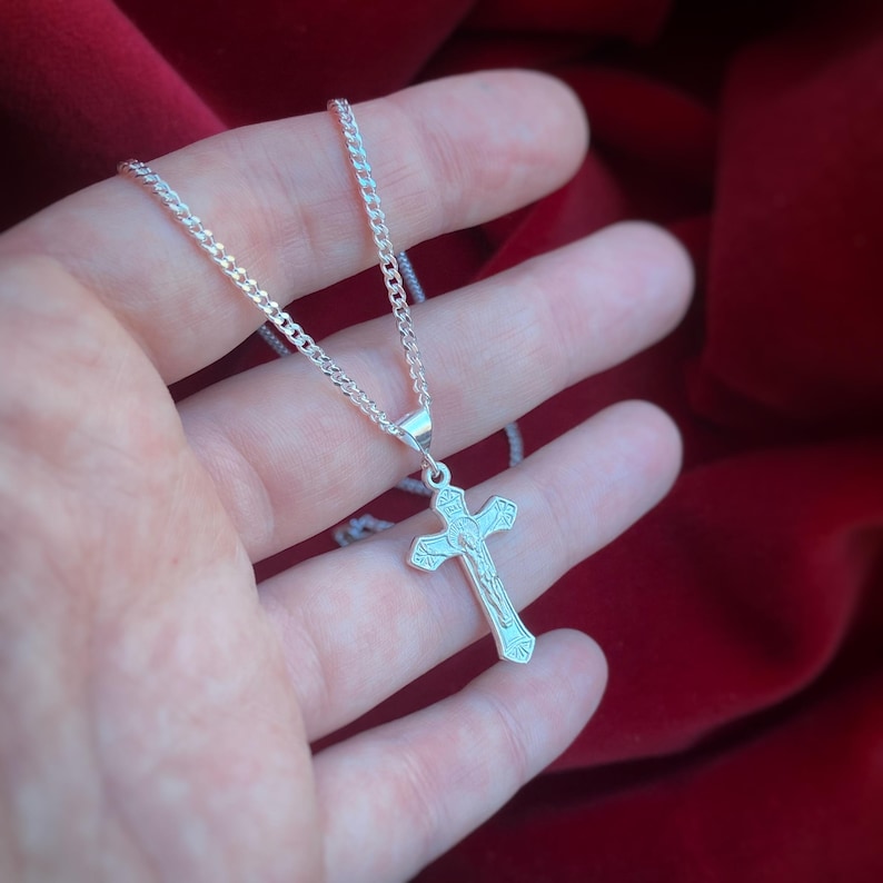 May include: A silver chain necklace with a silver cross pendant. The cross has a detailed design of a figure on it.