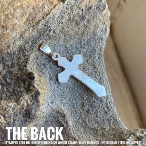May include: The back of a silver cross pendant with a sterling silver stamp. The stamp reads "STER" or "925" depending on which stamp is visible.