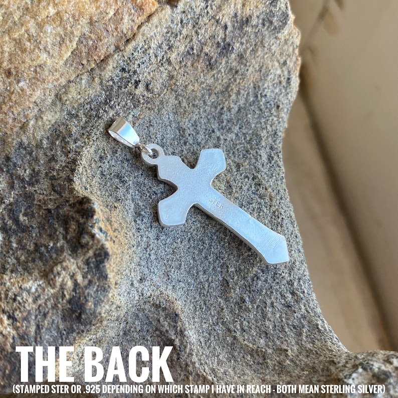 May include: The back of a silver cross pendant with a sterling silver stamp. The stamp reads "STER" or "925" depending on which stamp is visible.