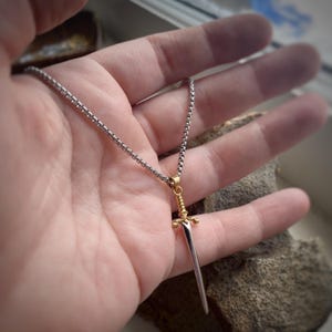 May include: A silver chain necklace with a gold sword pendant. The sword has a pointed tip and a decorative handle.