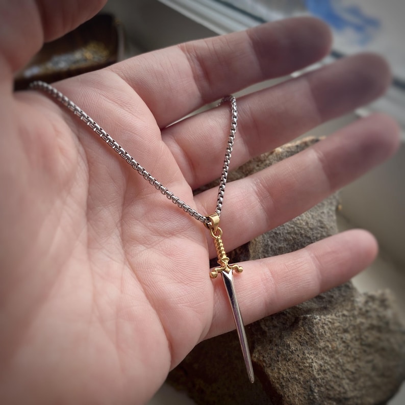 May include: A silver chain necklace with a gold sword pendant. The sword has a pointed tip and a decorative handle.