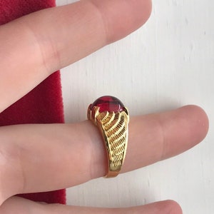 Peut inclure: Une bague en or avec une pierre précieuse rouge. La bague a une bande texturée.