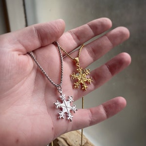 May include: Two silver snowflake necklaces, one with a gold snowflake pendant. The necklaces are on a hand, with the gold snowflake pendant on the left and the silver snowflake pendant on the right.