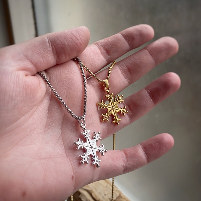 May include: Two silver snowflake necklaces, one with a gold snowflake pendant. The necklaces are on a hand, with the gold snowflake pendant on the left and the silver snowflake pendant on the right.
