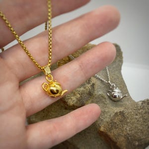 May include: A gold chain necklace with a small teapot pendant. The teapot is detailed with a spout, handle, and lid. The necklace is on a hand, and a silver chain necklace with a teapot pendant is visible in the background.