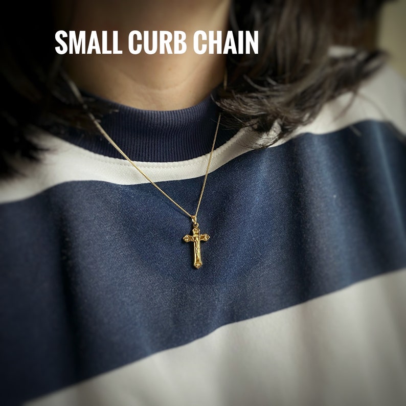 Pu&ograve; includere: Una collana a catena curb dorata con un piccolo ciondolo a croce dettagliato. La collana &egrave; indossata da una persona che indossa una camicia a righe blu e bianche. Il testo "SMALL CURB CHAIN" &egrave; visibile nella parte superiore dell'immagine.