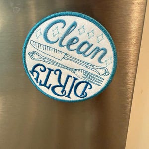 Op de afbeelding: Een ronde, geborduurde magneet met een witte achtergrond en een turquoise rand. De woorden "Clean" en "Dirty" zijn in turquoise geborduurd, samen met een mes- en vorkontwerp. De magneet is bevestigd op een roestvrijstalen oppervlak.