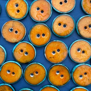 May include: A collection of orange ceramic buttons with two holes, set on a dark blue surface. Each button has a turquoise rim. The buttons are circular and have a glossy finish, creating a textured pattern.