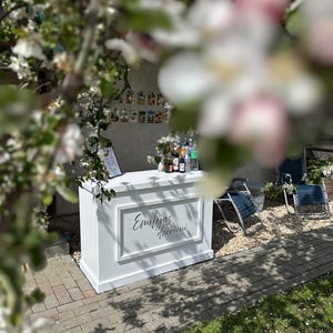 Op de afbeelding: Een witte buitenbar met de tekst "Emilijas dzērieni" in script. De bar staat op een stenen terras met groen gras en twee blauwe klapstoelen in de buurt. Diverse flessen en mixers staan op de bar.