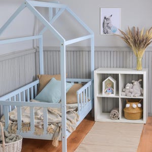 Peut inclure: Un lit cabane en bois bleu clair avec une tête de lit et un pied de lit à lattes. Le lit est orné de coussins et d'une couverture. Une étagère blanche contient des jouets et un panier tressé. Une impression de zèbre encadrée est accrochée au mur.